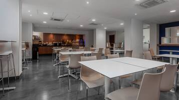 A modern dining area with white tables and chairs, a kitchen in the background, and a blue accent wall.
