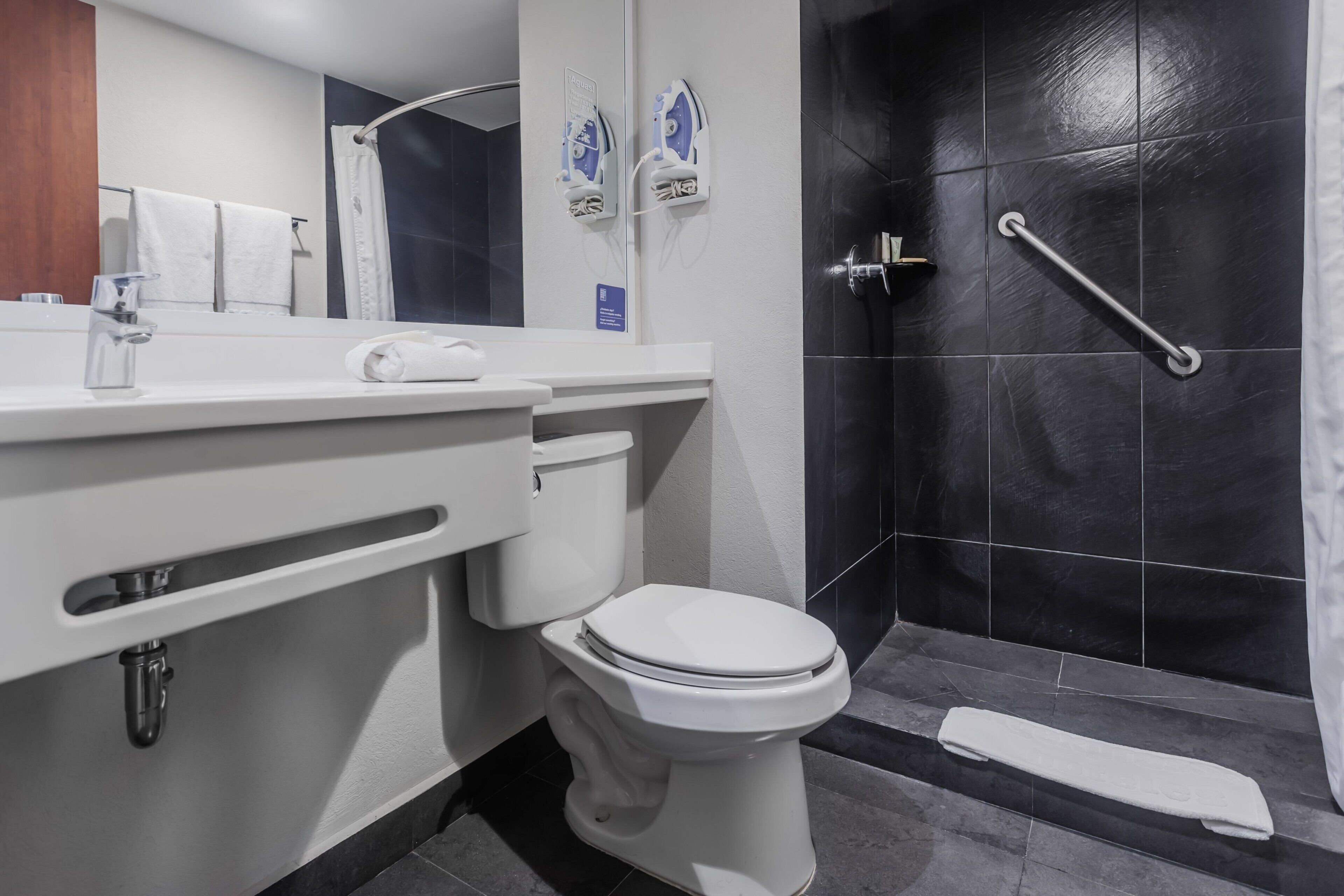 Un baño moderno con inodoro blanco, lavamanos con espejo y zona de ducha con barra de apoyo.