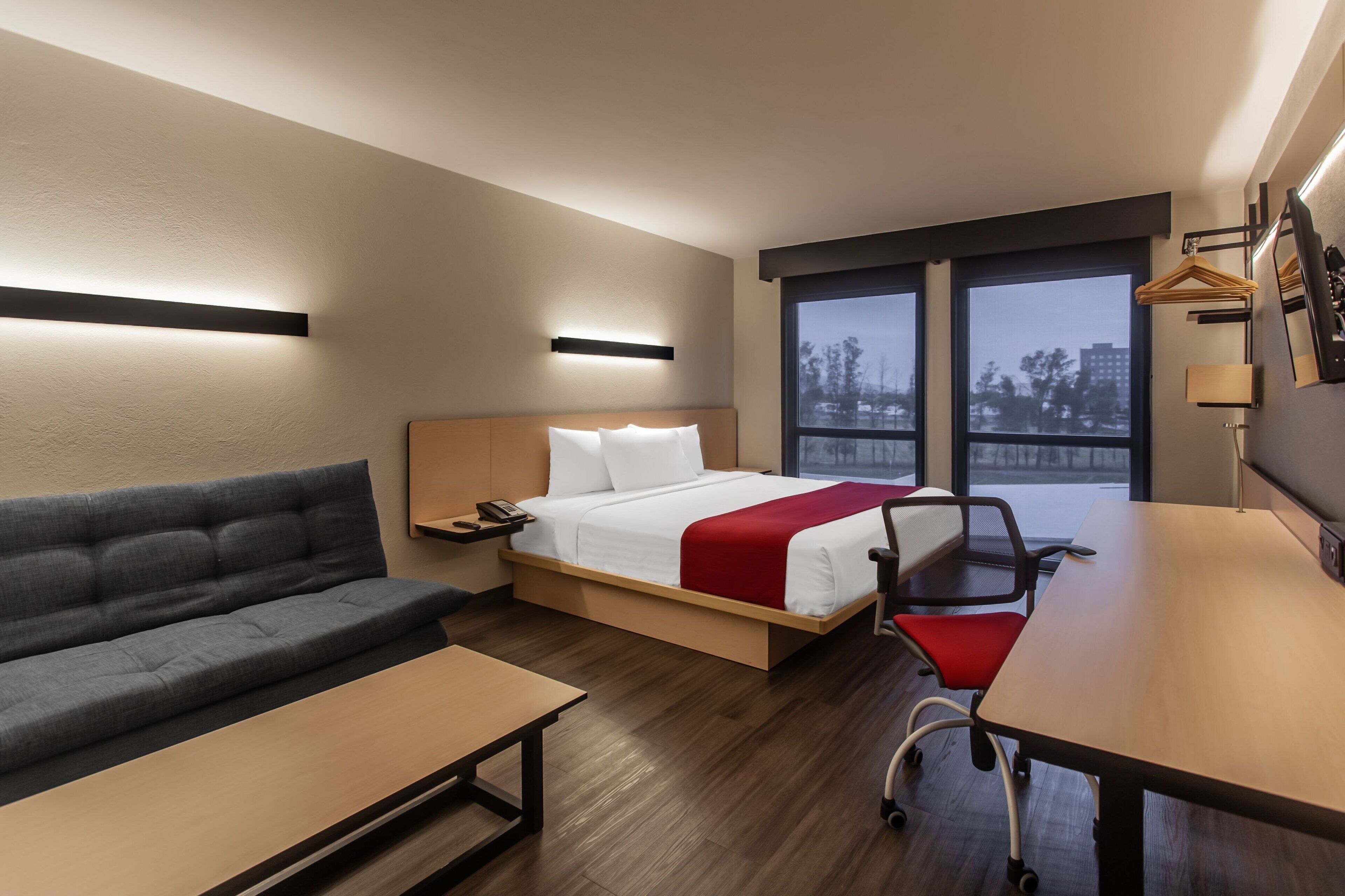 A hotel room with a bed, a sofa, a desk, and a view of the city through large windows.