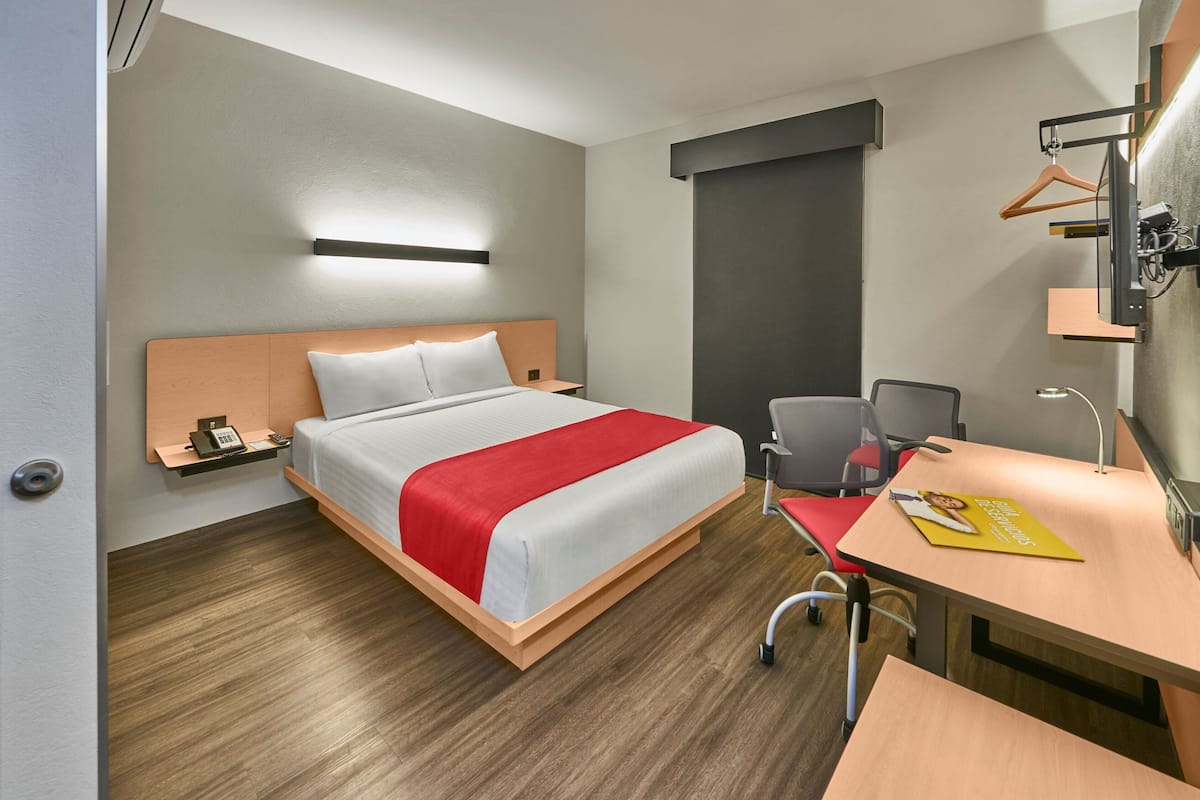 A hotel room with a bed, a desk, a chair, a bedside table, a hanging light, and a wardrobe.