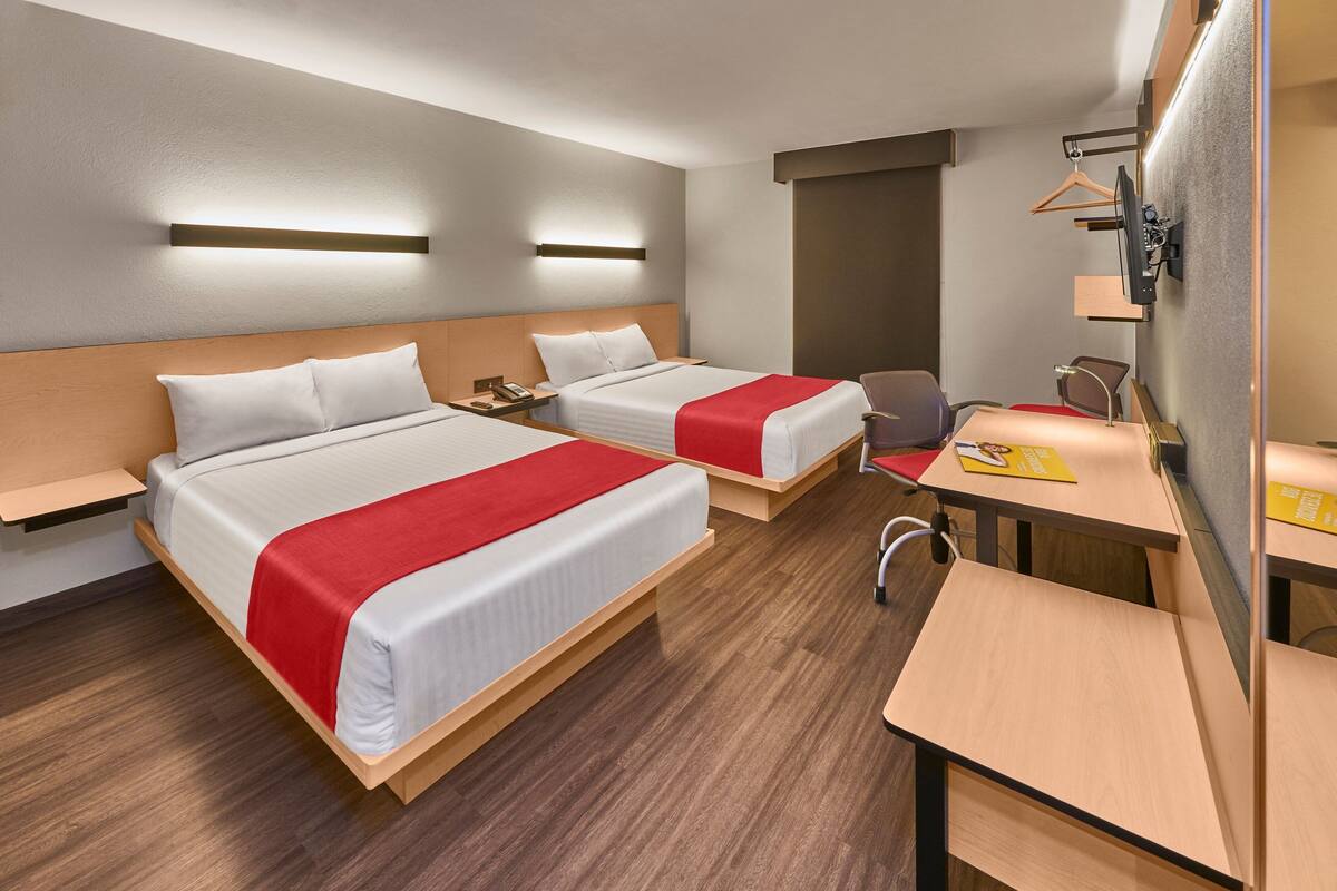 A hotel room with two beds, a desk, a chair, a desk lamp, a telephone, and a wall-mounted TV.