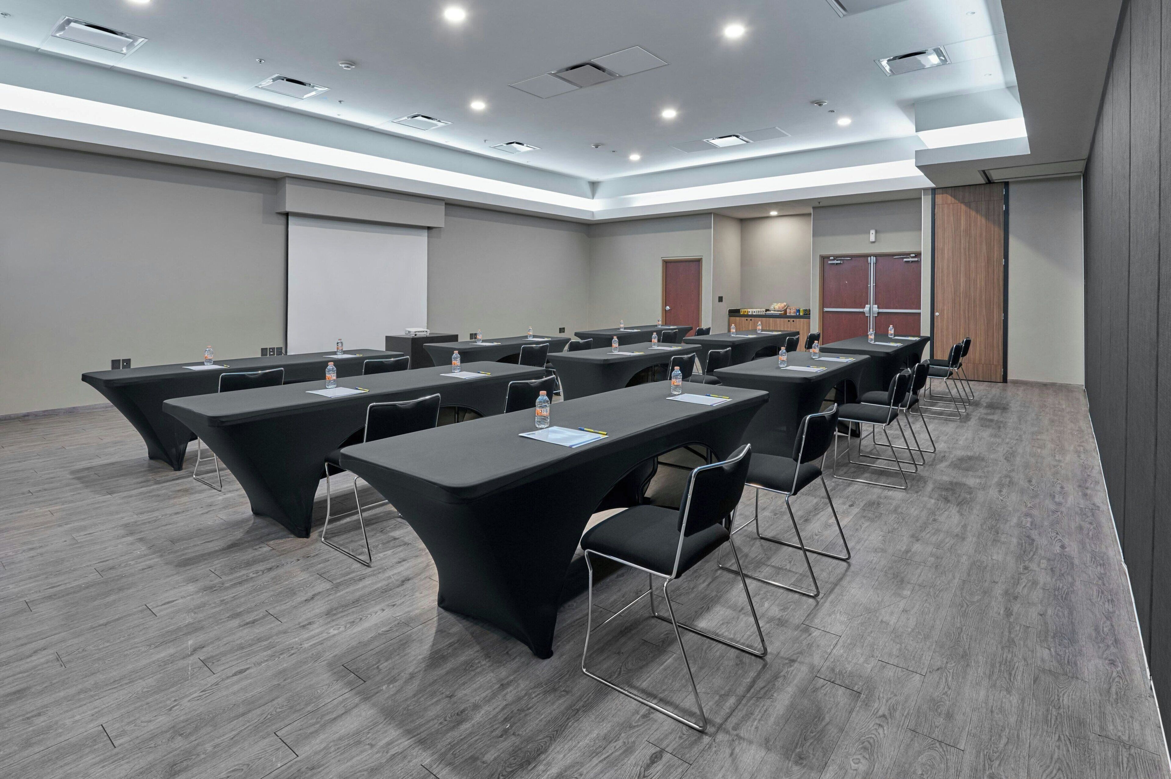Un salón de conferencias con varias mesas y sillas, equipado con botellas de agua y documentos, en un edificio moderno con piso gris e iluminación empotrada.
