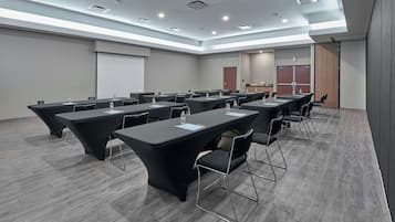 Un salón de conferencias con varias mesas y sillas, equipado con botellas de agua y documentos, en un edificio moderno con piso gris e iluminación empotrada.