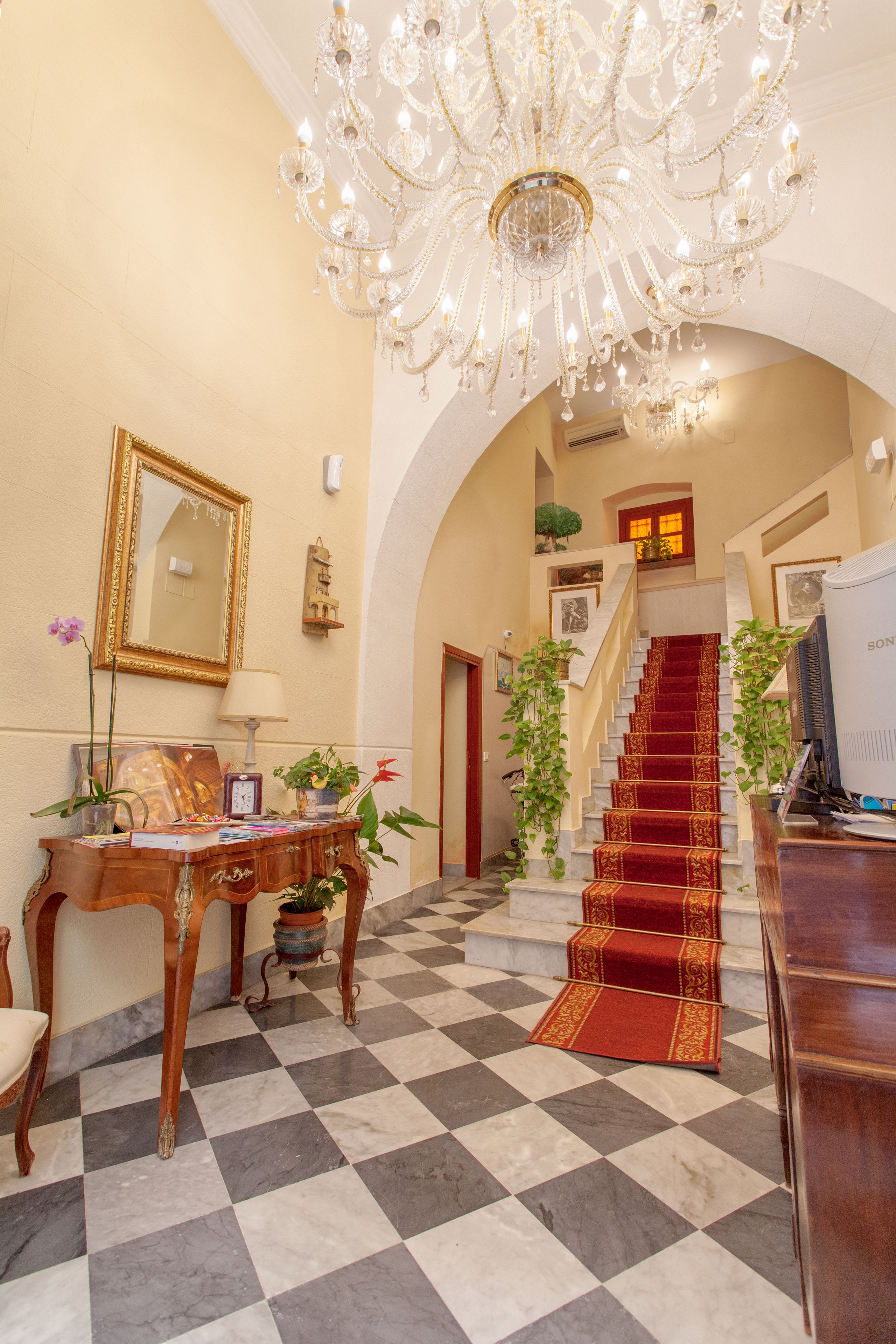 A grand lobby with a checkered floor, a chandelier, a staircase, and a reception desk.