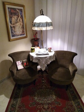 A room with two armchairs, a small table with a white lace tablecloth, a floor lamp, a framed picture on the wall, and a red patterned rug. - Al Piazza Marina B&B (Palermo)