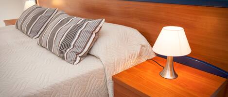 Una cama bien hecha con un almohadón a rayas, una lámpara de noche y una mesita de noche de madera con un cajón.