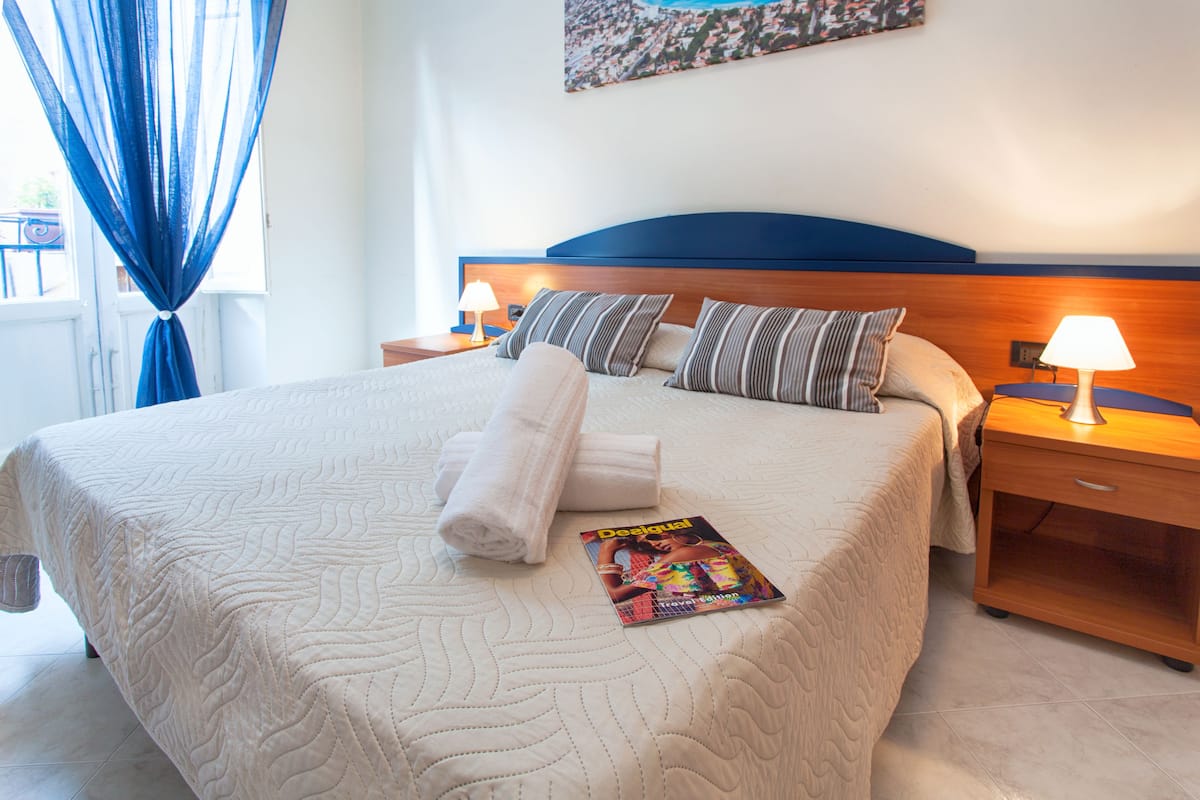 A neatly made bed with a quilted cover, pillows, and a magazine on it. There are two bedside tables with lamps, a window with blue curtains, and a wall-mounted picture.