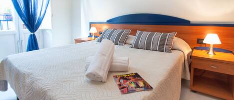 A neatly made bed with a quilted cover, pillows, and a magazine on it. There are two bedside tables with lamps, a window with blue curtains, and a wall-mounted picture.