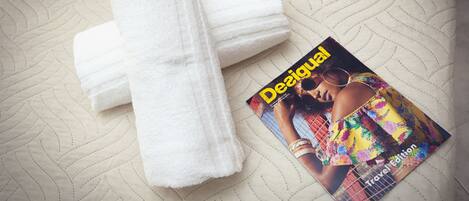 Toalha branca dobrada e uma revista intitulada "Desigual Travel Edition" sobre um cobreleito texturizado.