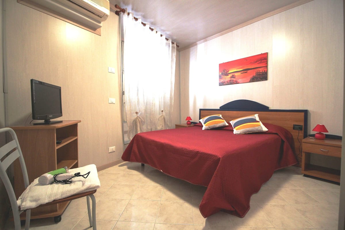 A bedroom with a bed, a chair, a desk, a TV, and a painting on the wall.
