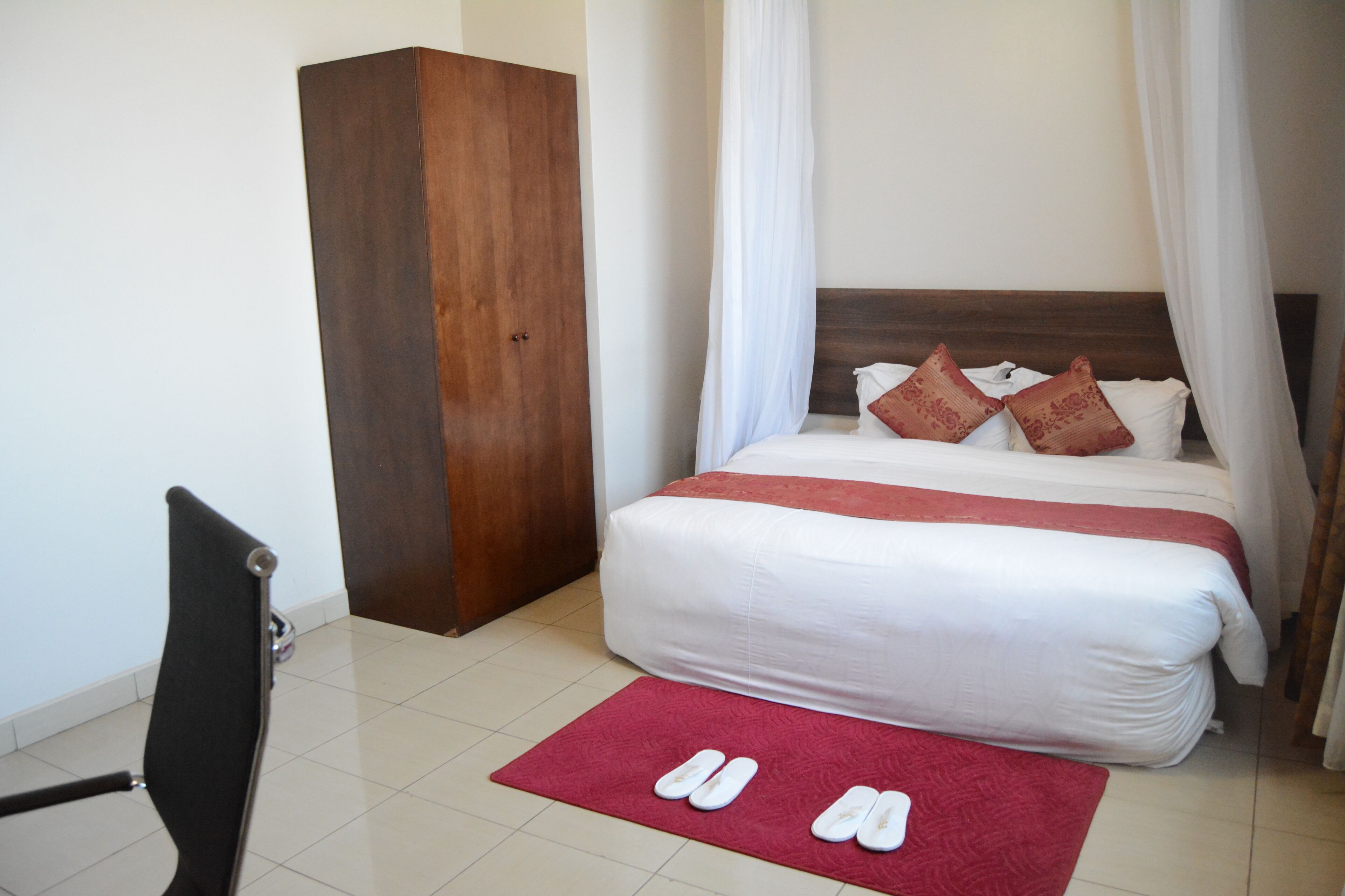 A hotel room with a bed, a wardrobe, a chair, and a red rug with slippers.