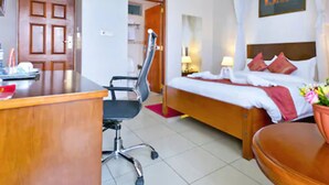 Una habitación de hotel con un escritorio de madera, una silla de oficina negra, una cama con almohadas rojas, una mesita de noche con lámpara y un televisor montado en la pared.