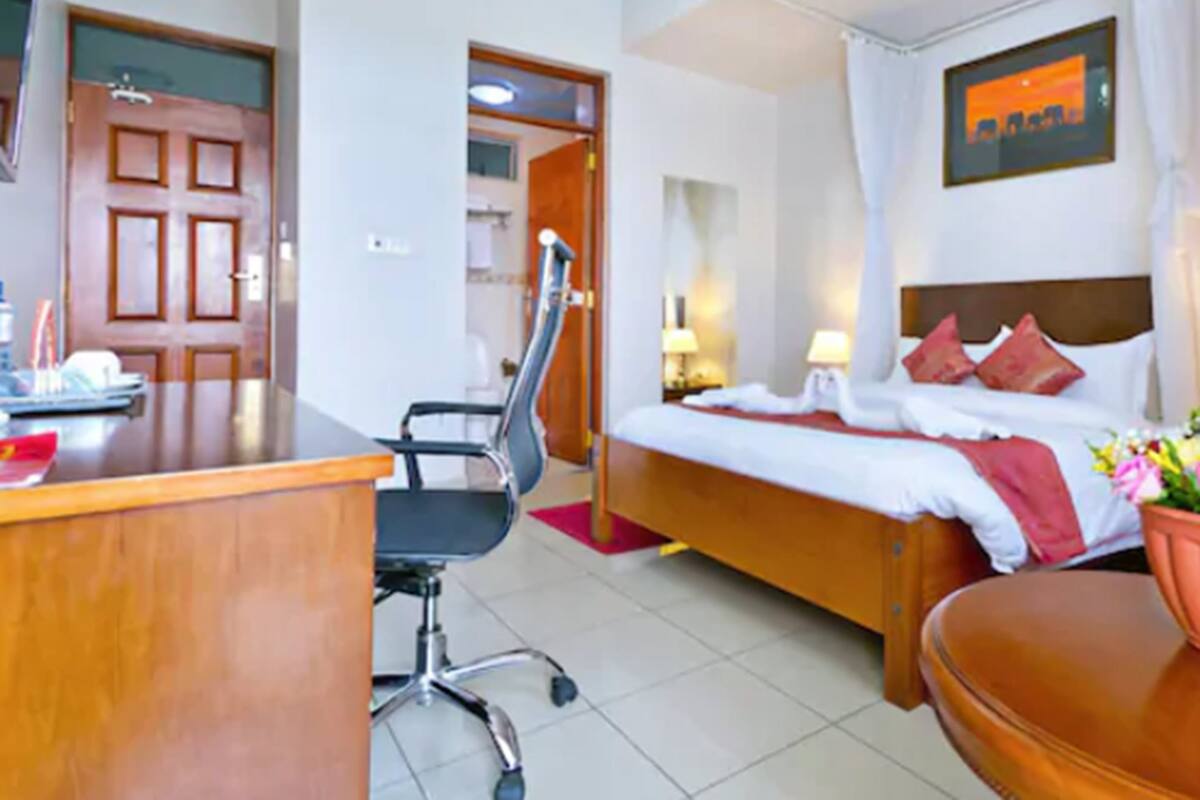 Ein Hotelzimmer mit Holztisch, schwarzem Bürostuhl, einem Bett mit roten Kissen, einem Nachttisch mit Lampe und einem Wand-Fernseher.