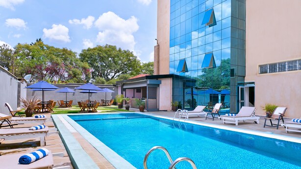 Piscina con sillones de descanso y sombrillas, rodeada por un edificio de fachada de vidrio.