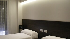Ein Hotelzimmer mit zwei Einzelbetten, einem dunklen Kopfteil, einem Fenster mit durchsichtigen Vorhängen und einem Nachttisch.