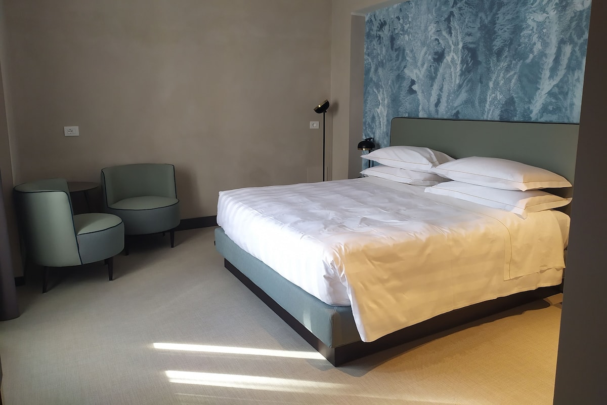 A hotel room with a bed, two chairs, a floor lamp, and a large abstract painting on the wall.