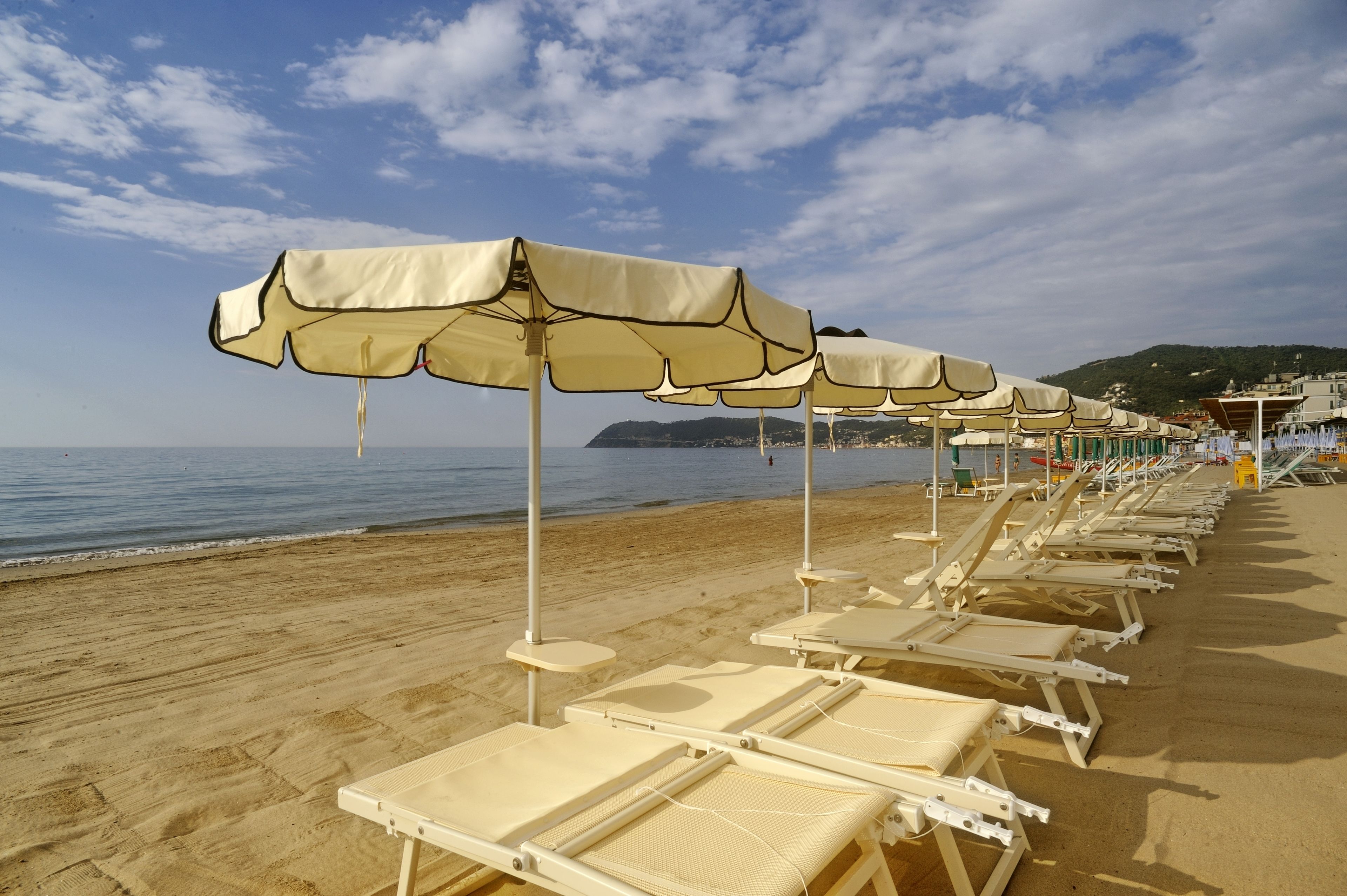 Una playa con filas de tumbonas y sombrillas, una orilla arenosa y vistas al mar y colinas lejanas.