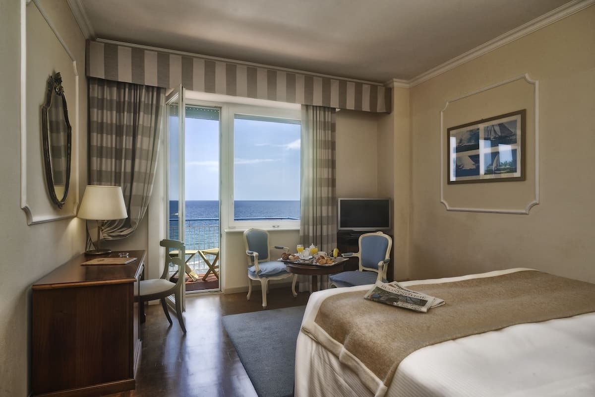 A hotel room with a bed, a desk with a lamp, a chair, a small table with breakfast, a TV, and a window with a view of the sea.