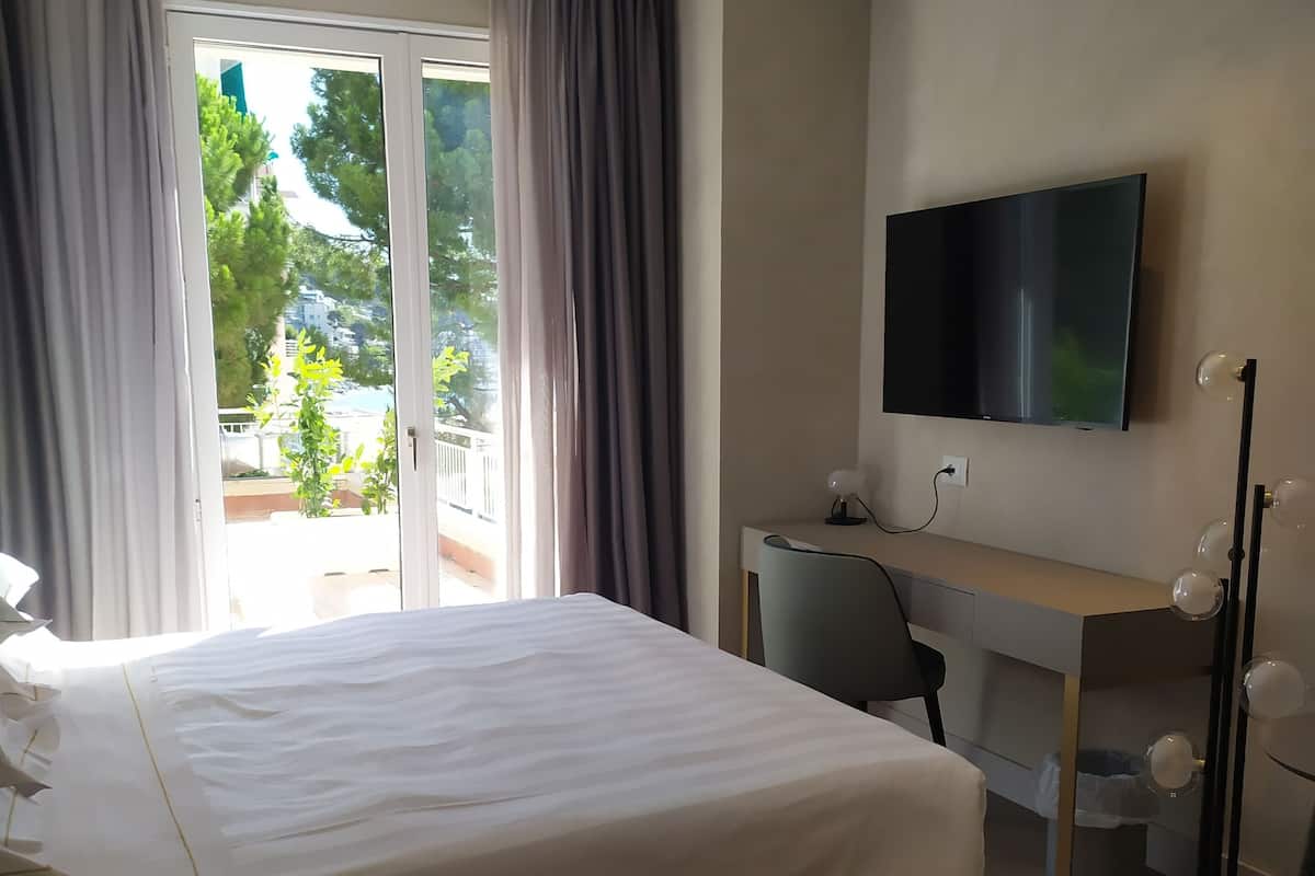 A hotel room with a large bed, a desk, a TV, and a view of greenery outside.