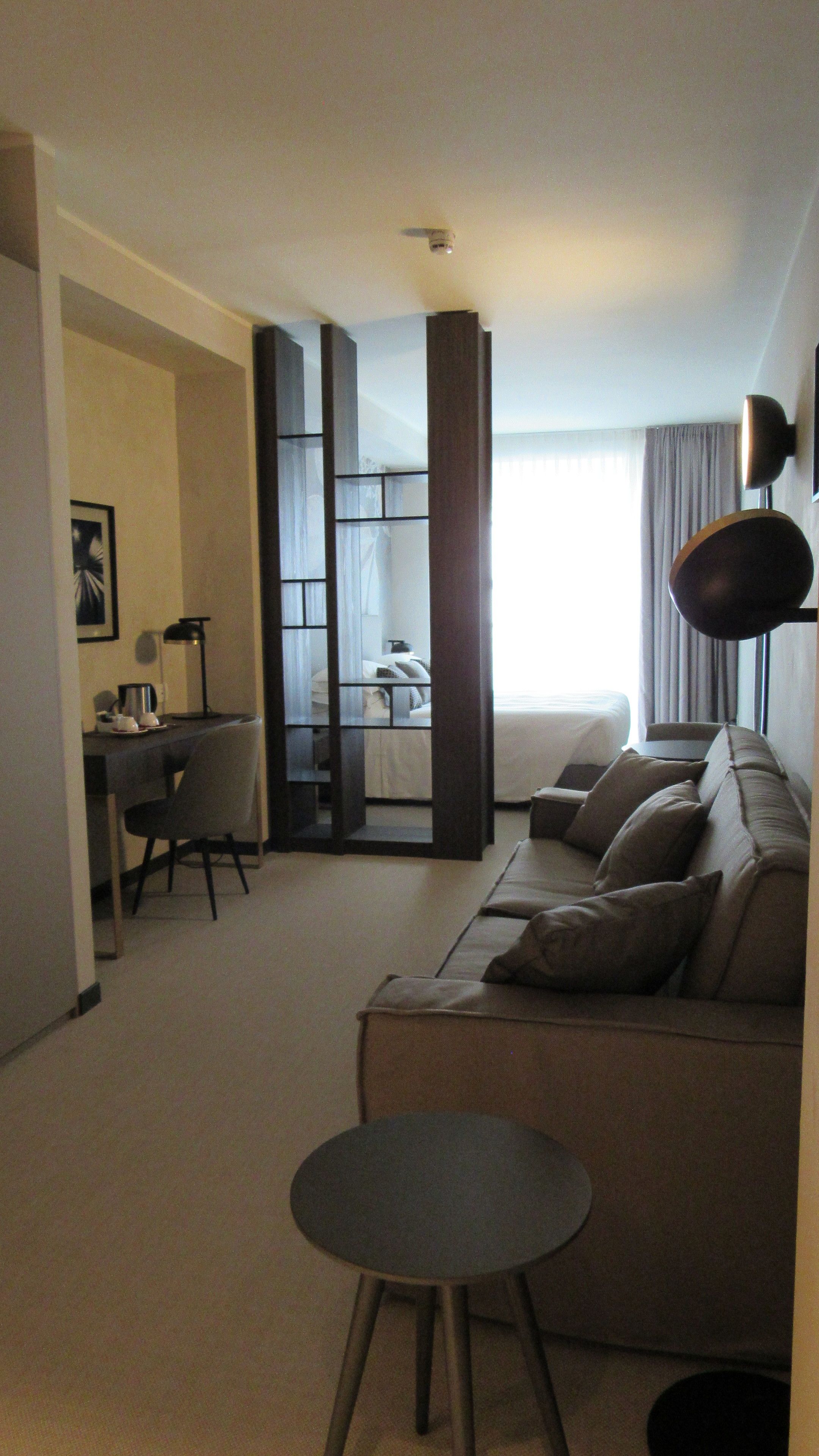 A modern living room with a sofa, a round coffee table, and a desk with a chair. There is a large window with curtains, a lamp, and a framed picture on the wall.