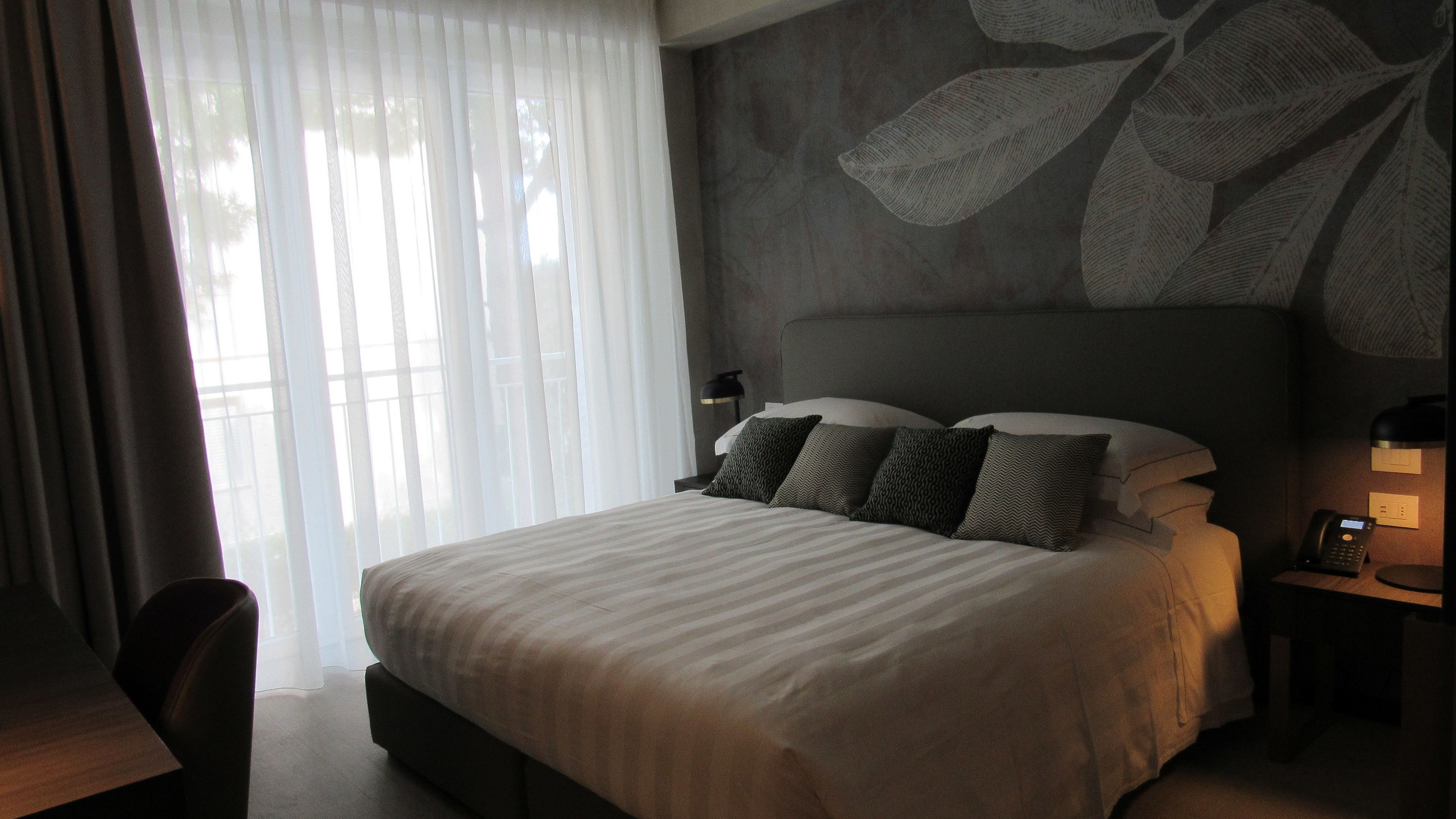A hotel room with a large bed, a desk, and a chair. There is a window with sheer curtains and a wall with a leaf pattern.