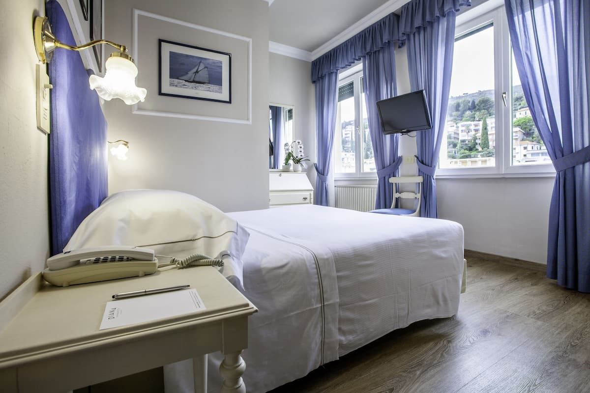 A hotel room with a bed, bedside table with a telephone and pen, a mirror, a TV, and a window with curtains.