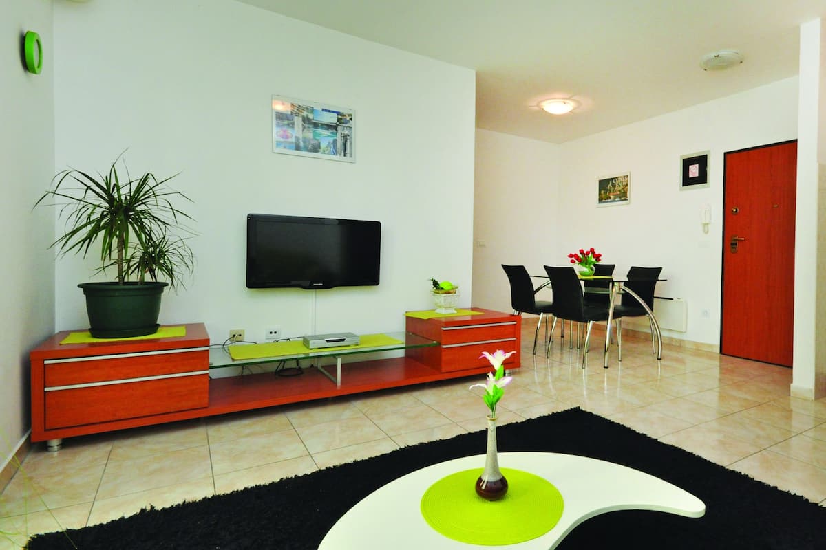 A modern living room with a flat-screen TV, a dining area with chairs, a red door, and a potted plant.