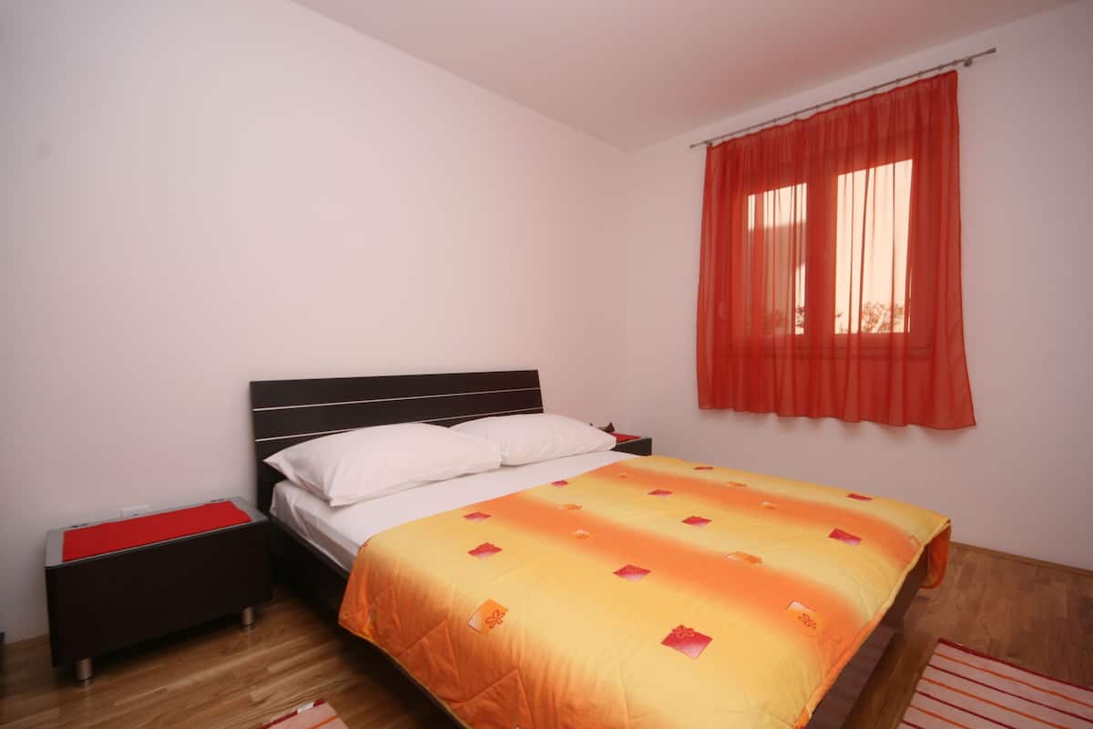 A bedroom with a bed, a nightstand, a wooden floor, and a window with red curtains.