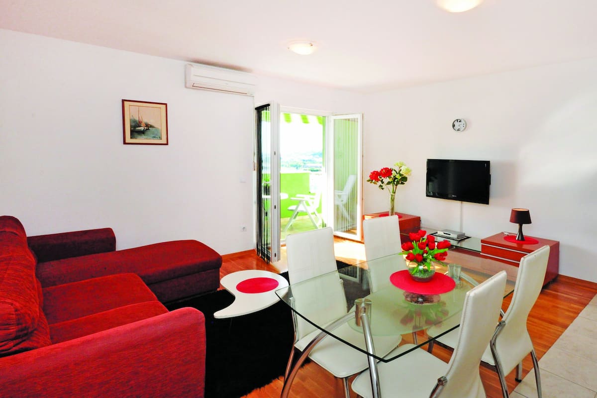 A modern living room with red sofas, a glass dining table with chairs, a TV, and a view through a glass door.