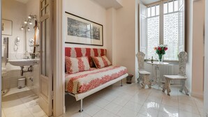 Une chambre à coucher avec un lit, une fenêtre ornée de rideaux en dentelle, un radiateur et une petite table sur laquelle est posé un vase de fleurs.
