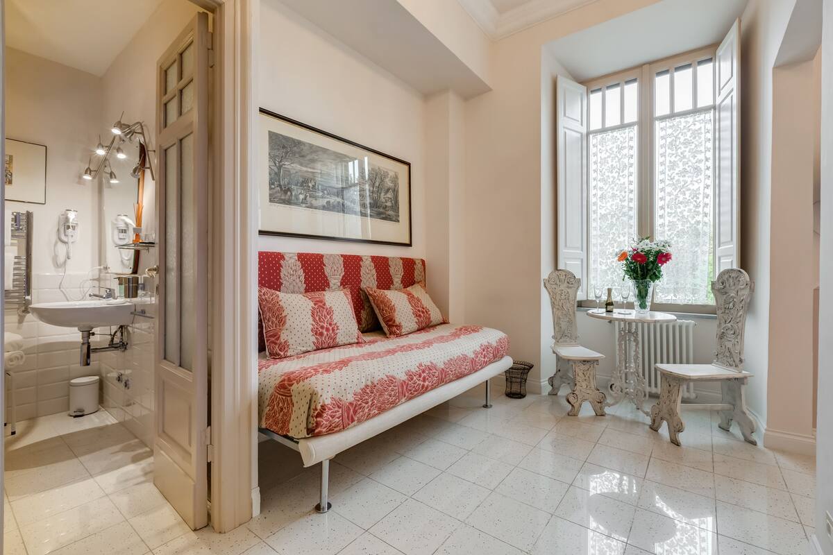 Un dormitorio con cama, una ventana con cortinas de encaje, un radiador y una pequeña mesa con un florero.