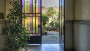 Une porte ouverte, ornée de vitraux, qui donne sur une cour intérieure agrémentée de plantes en pot et bordée d’un mur de briques.