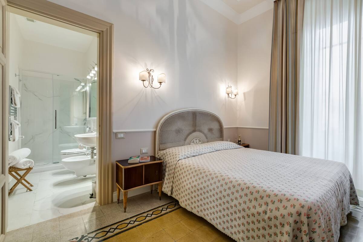Un dormitorio con cama, mesitas de noche, un tocador, un espejo y un baño visible a través de una puerta abierta.