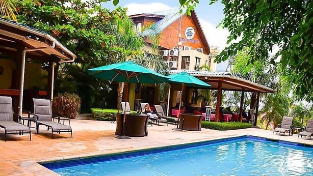 A swimming pool with lounge chairs and umbrellas, surrounded by greenery and a building with a tiled roof. - Elite Hotel (Port-au-Prince)