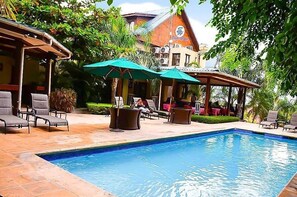 Piscina con sillones de descanso y sombrillas, rodeada de vegetación y un edificio con techo de tejas.