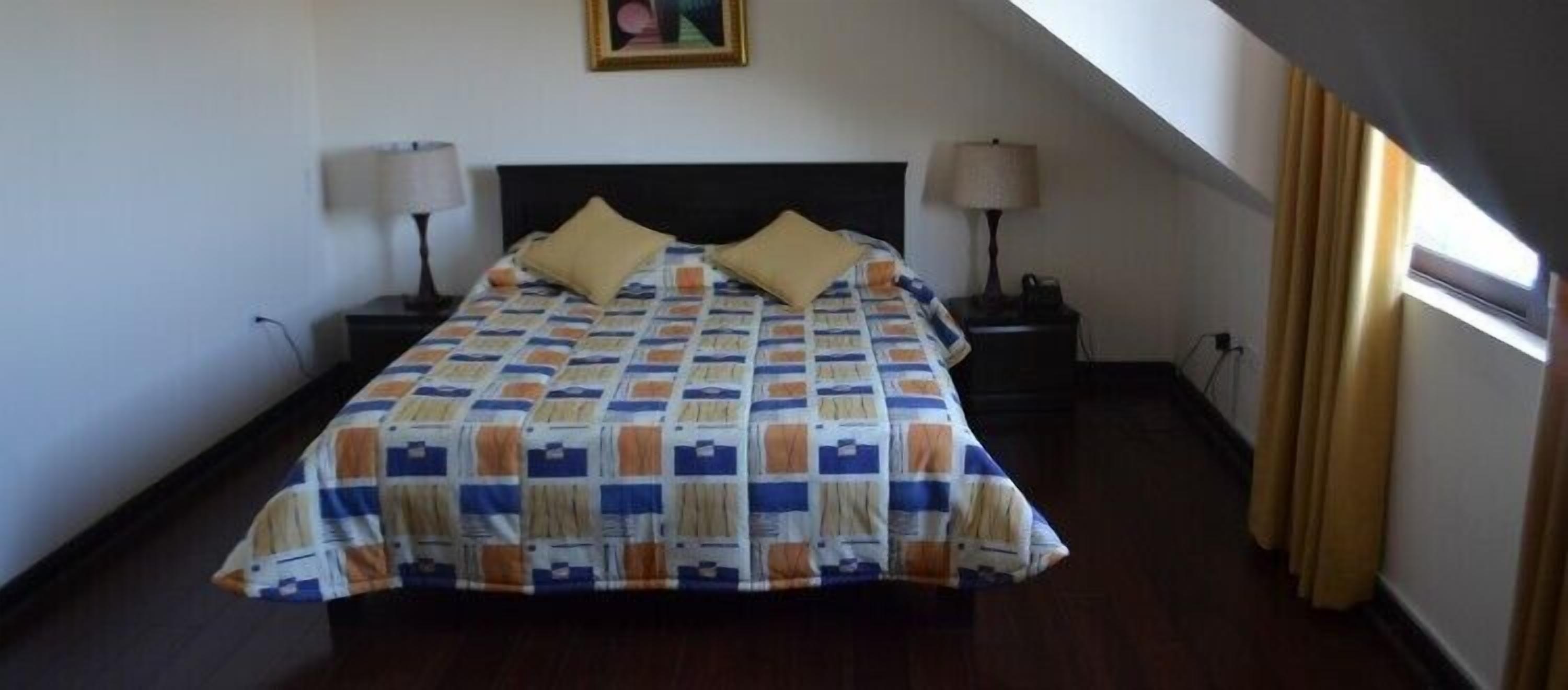 A bedroom with a bed, two bedside tables, a nightstand, a window with curtains, and a painting on the wall.