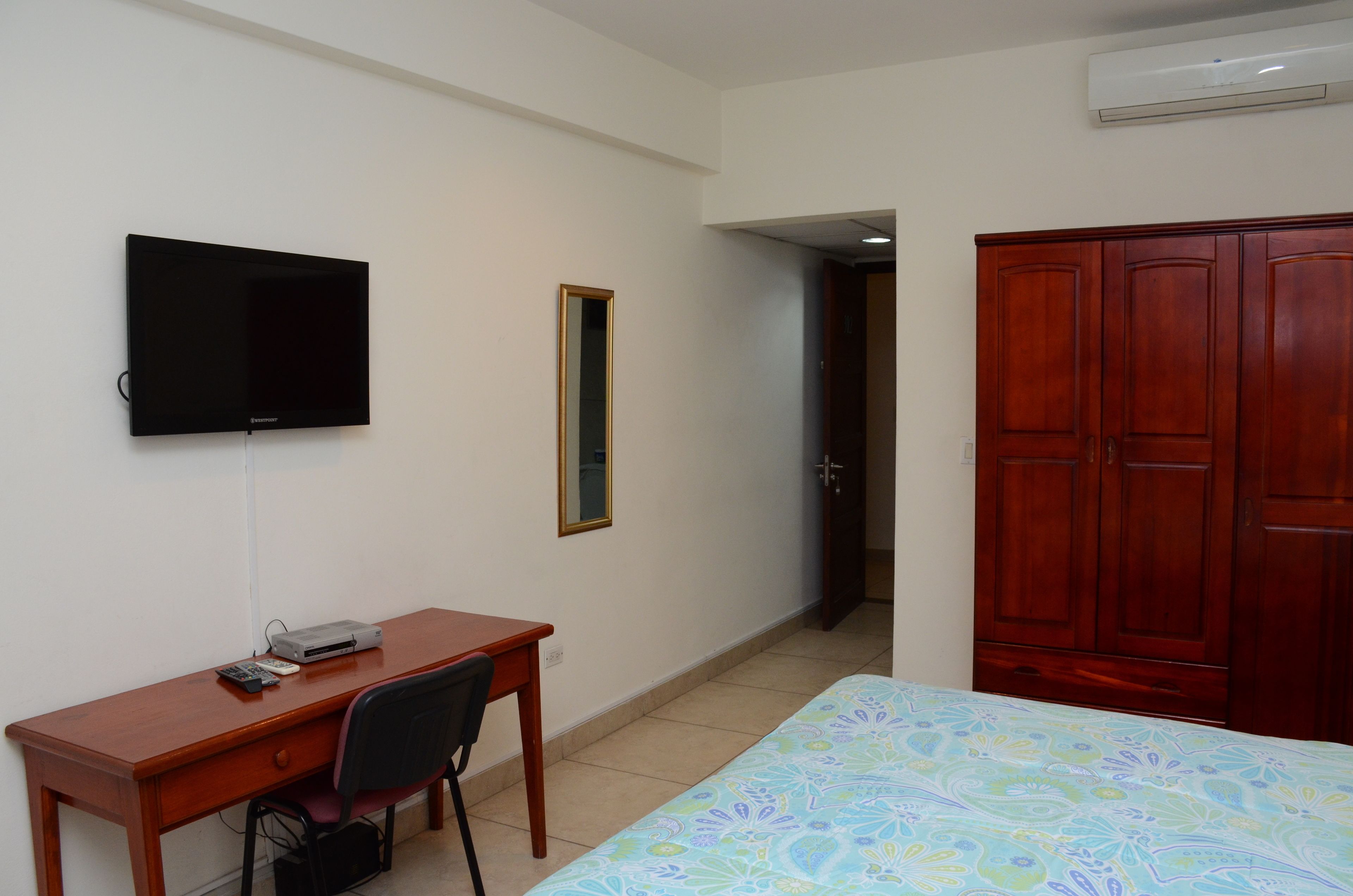 A room with a bed, a desk with a chair, a television, a wardrobe, and a mirror.