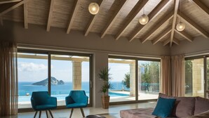 A spacious living room with a large window overlooking a pool and the sea. There are two blue chairs and a brown sofa with cushions. - Avra Luxury Villa (Zakynthos)