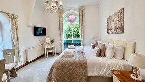 A bedroom with a large bed, a bedside table with a lamp, a TV on the wall, and a window with curtains. - The Chapel Guest House (St Austell)