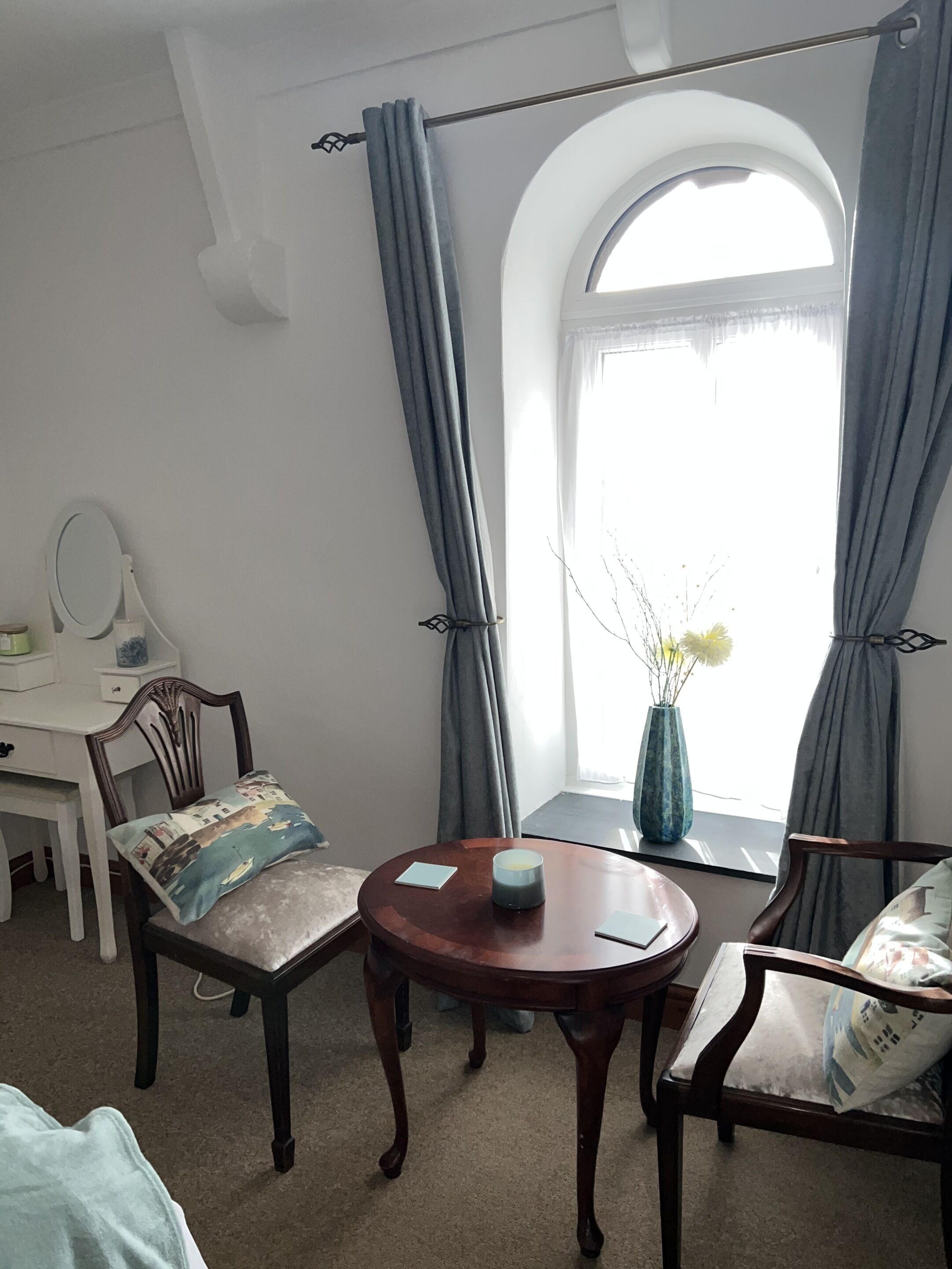 A room with a round wooden table, two chairs, a desk with a mirror, and a window with curtains.