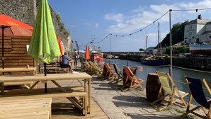 A wooden deck with tables and chairs, a stone wall, and a river with boats. - The Chapel Guest House (St Austell)