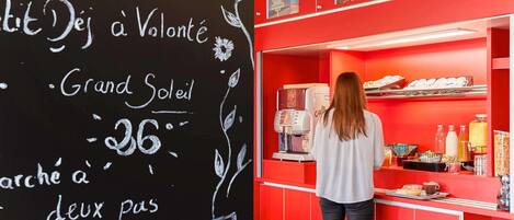 A woman at a red counter with a coffee machine and various food items.