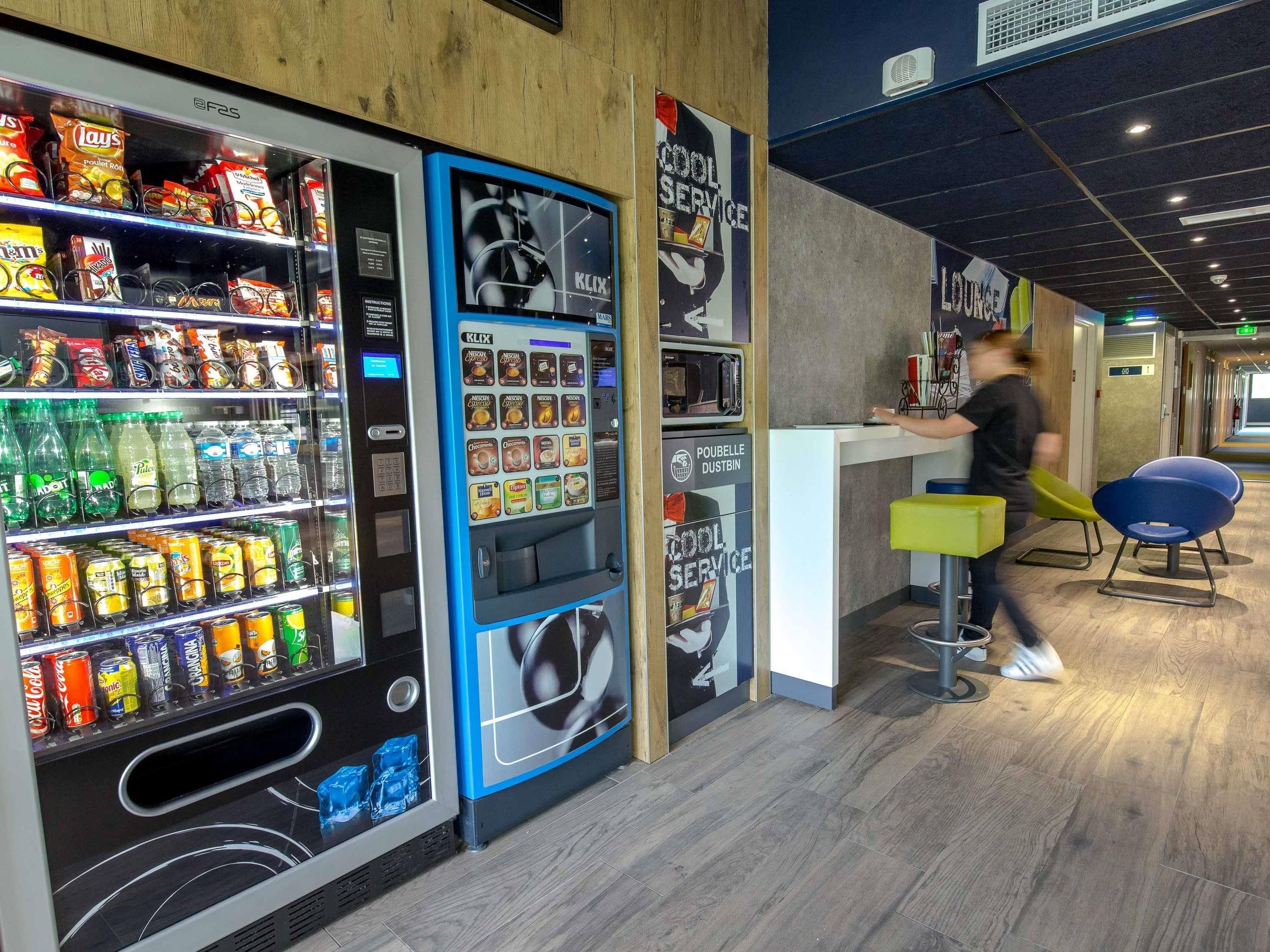 Máquina expendedora con aperitivos y bebidas, mostrador con una persona y área de descanso con sillas.
