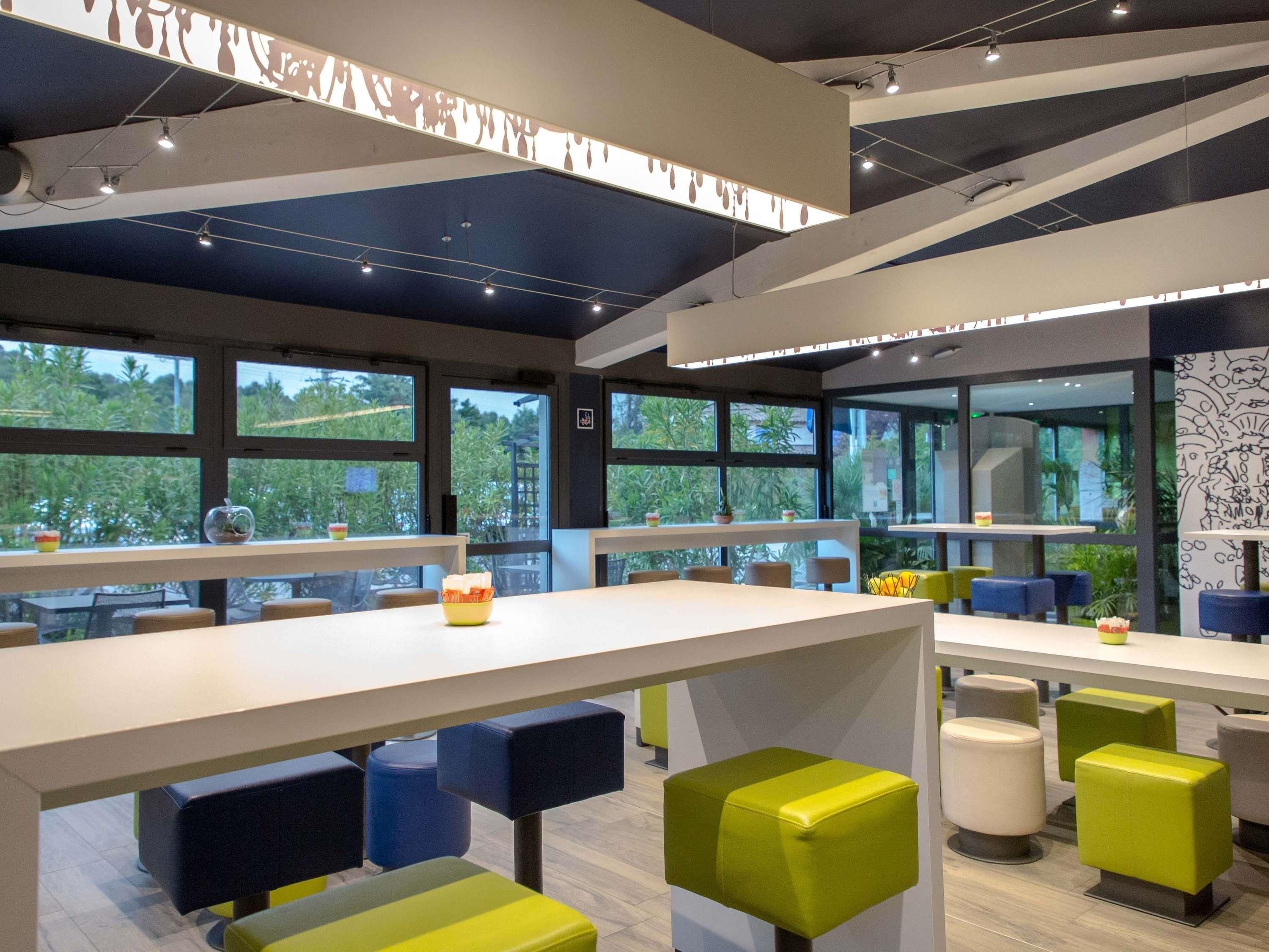 A modern indoor space with tables and chairs, large windows, and a ceiling with exposed beams and lighting fixtures.