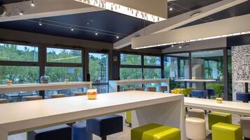 A modern indoor space with tables and chairs, large windows, and a ceiling with exposed beams and lighting fixtures.