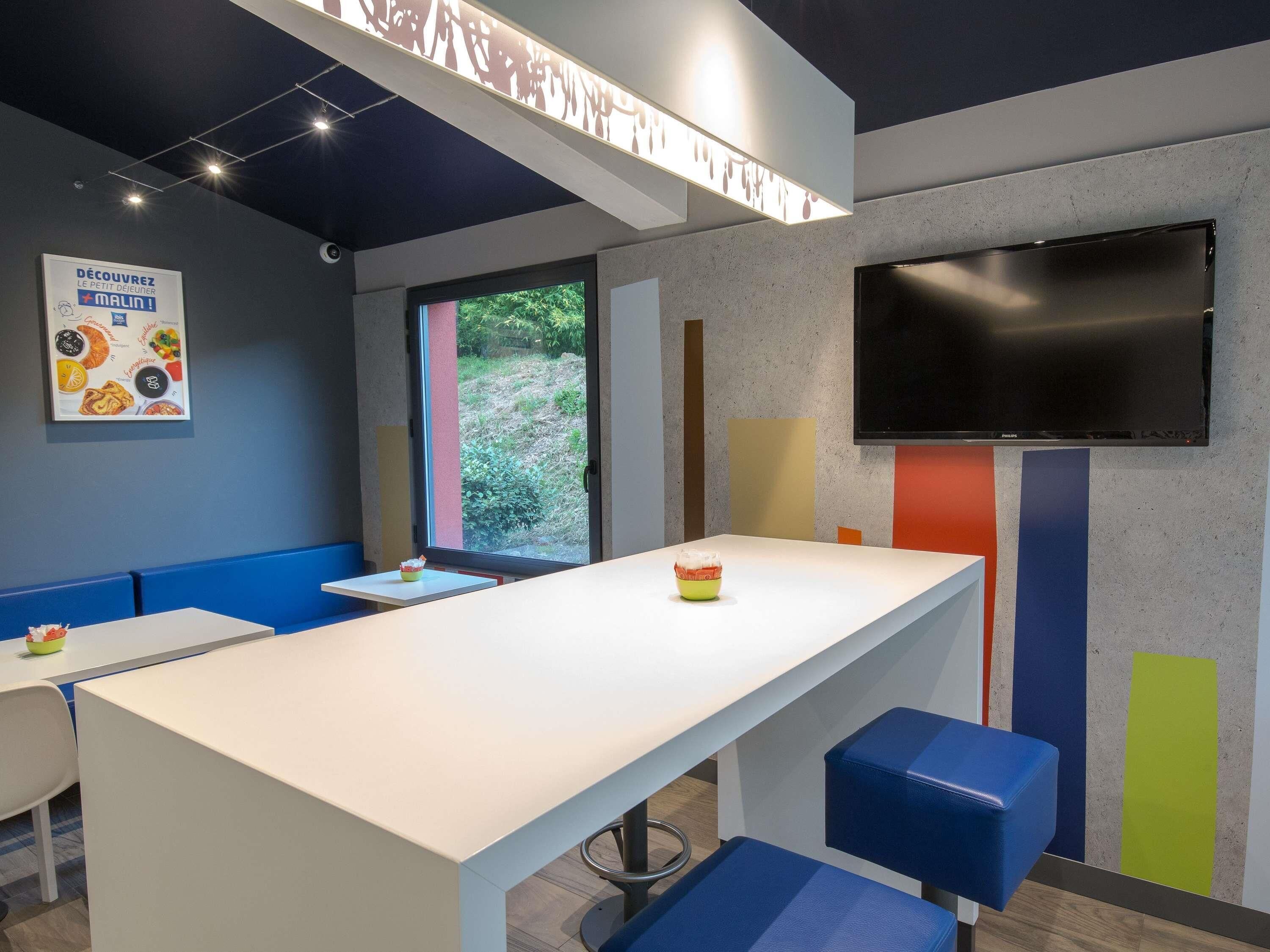 A modern interior with a long white counter, blue seating, a large window, and a flat-screen TV on the wall.