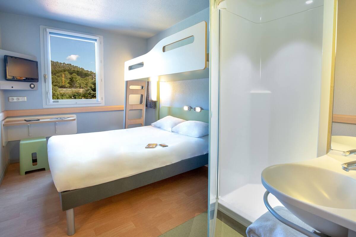 Quarto de hotel moderno com beliche, uma cama de solteiro, banheiro com chuveiro em box de vidro, pia e TV fixada na parede.