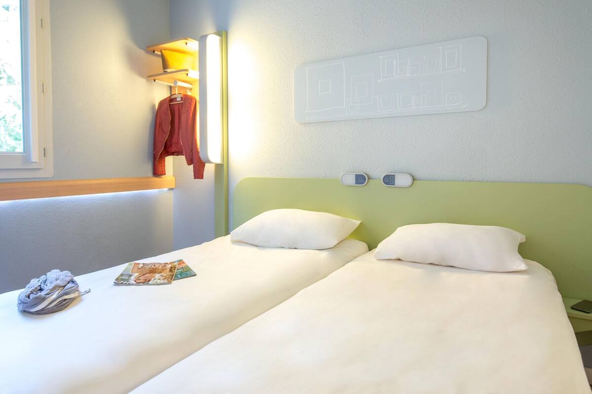 Uma cama de casal com roupa de cama branca, uma mesinha de cabeceira com telefone, e uma luminária fixada na parede.
