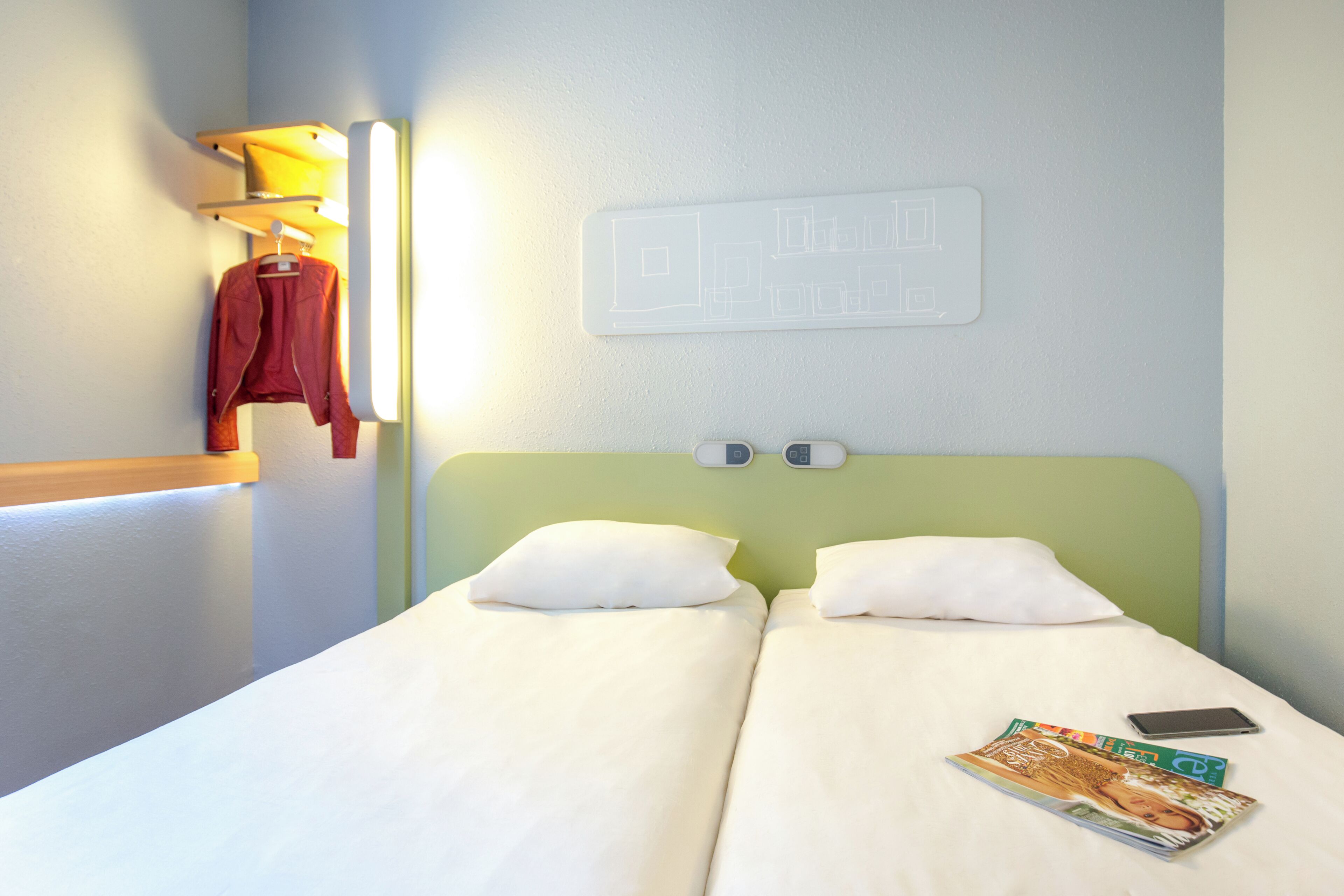 Une chambre d’hôtel avec un lit, des oreillers, un magazine, un téléphone intelligent, une lampe fixée au mur et un porte-manteau avec une veste.