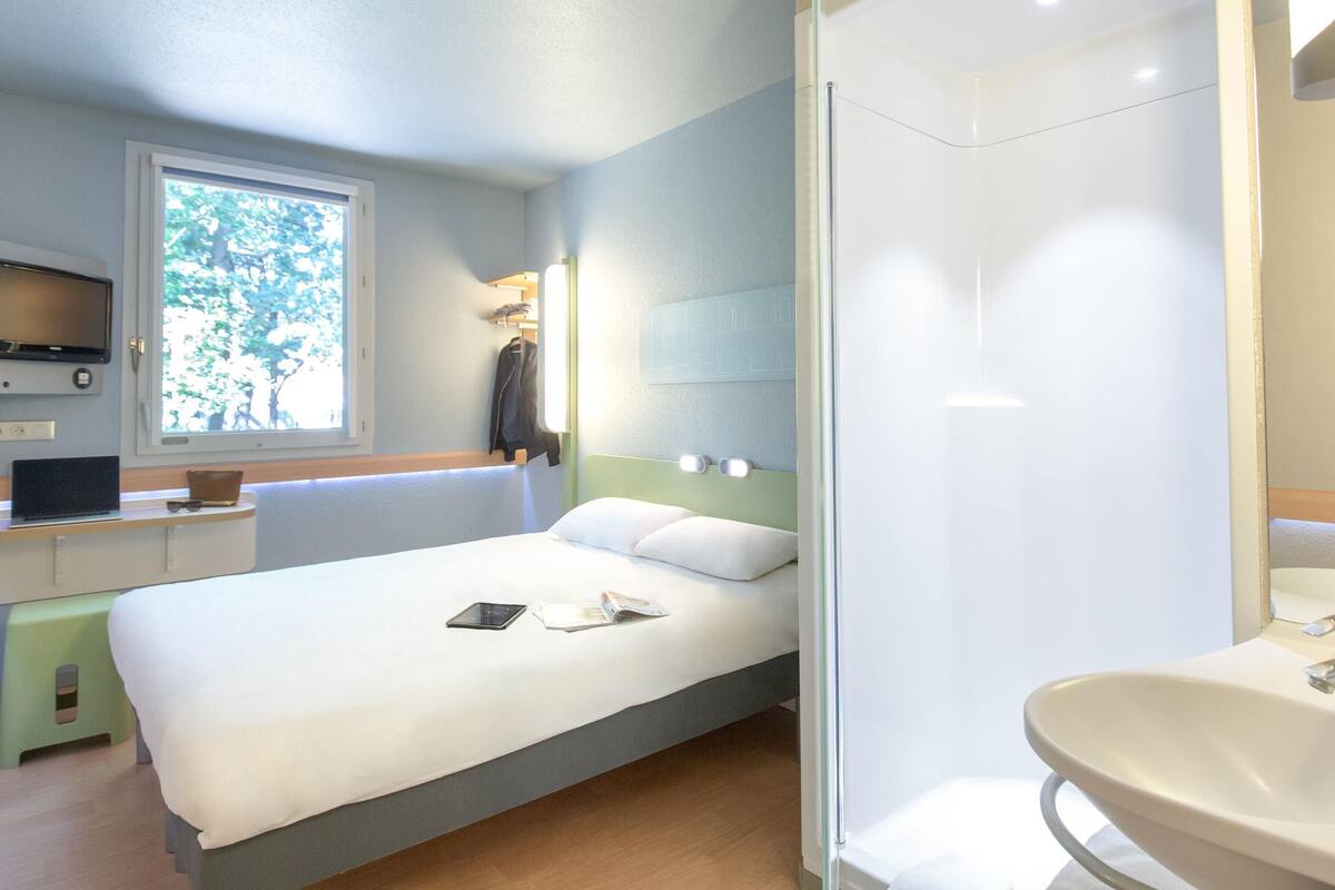 Quarto de hotel moderno com cama, mesa de cabeceira, televisão e banheiro com pia e espelho.